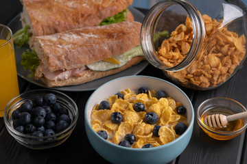 Breakfast - tasty corn flakes with berry and honey, sandwich with salad and meat, baguette with cheese and tomato. Kitchen. Black table background