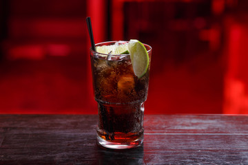 Closeup of a Cuba Libre cocktail in short glass, gin, standing on the bar counter, isolated on a red light background. horizontal view.