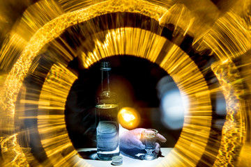 A bottle of alcohol is on a table. A man's hand is holding a shot glass. Bright lights symbolize drunkenness. Concept of alcoholism.