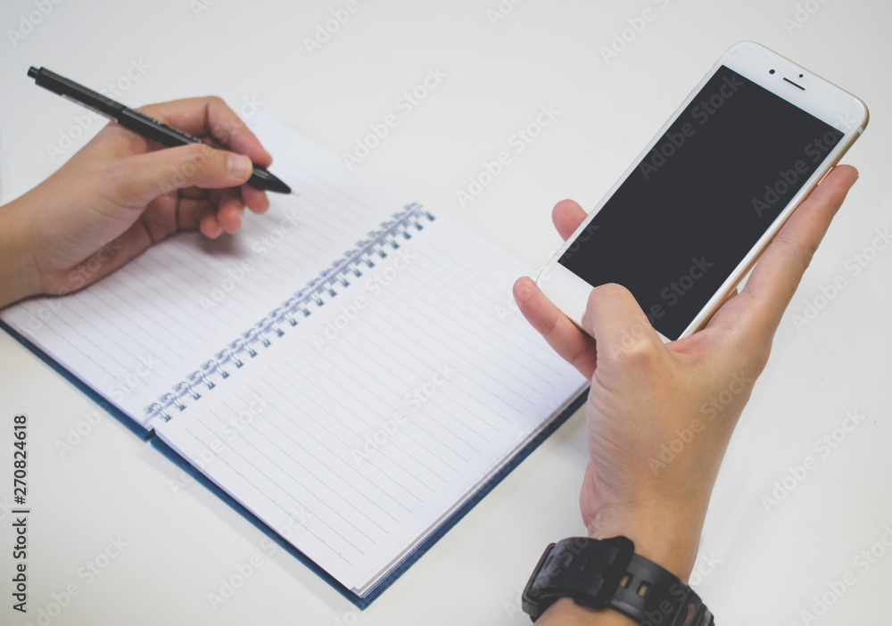 Poster close up image. a hands with pen writing on notebook and using smartphone at the same time.