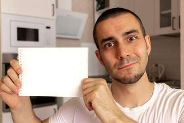 Young man showing blank paper notepad with space for text. Kitchen background blurred.