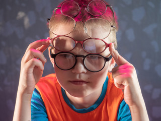 Fototapeta na wymiar Photo of funny boy, blonde kid with lot of pairs of glasses on his face. Child is happy at home on vacation.