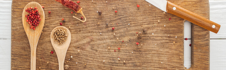 panoramic shot of knife, spoons, scattered coriander and pink peppercorn, dried chili pepper on wooden chopping board