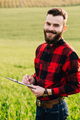 Young handsome bearded farmer with folder standing in green wheat field in early summer