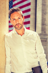Naklejka premium Thinking outside. Young European Businessman with beard, wearing white shirt, holding briefcase, standing outside office building on street in New York City, under sunshine, looking, thinking. .