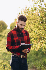 Young handsome bearded farmer with folder standing in green fruit garden in early spring