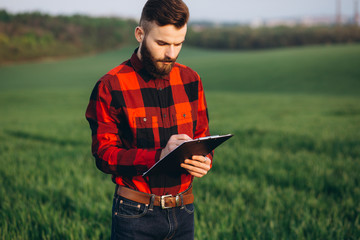 Young handsome bearded farmer with folder standing in wheat field in early summer