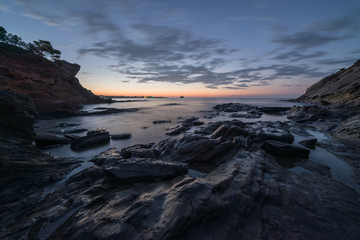 Rocks among the seashore at sunrise. There are cliffs at the sides under a cloudy sky.