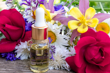 Essential oil in a glass bottle near wildflowers on wooden background.