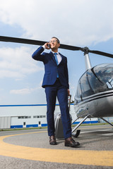businessman in formal wear with suitcase talking on smartphone near helicopter