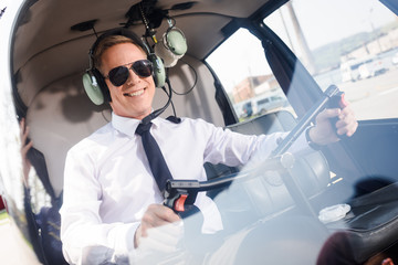 smiling Pilot in sunglasses and headset with microphone sitting in helicopter cabin and holding...