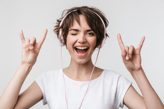 Image of attractive woman in basic t-shirt showing rock sign while listening to music with headphones