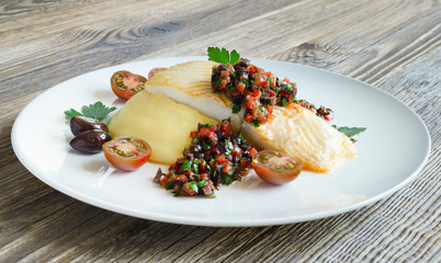 fried halibut fillet with mashed potato and tomato-olives salsa on white plate on wooden background