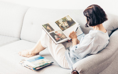 Middle-aged brunette woman on the gray sofa looking at album with paintings by artists.