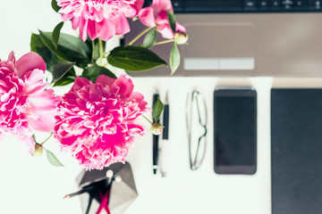 bouquet of pink peonies, laptop, smartphone, pens, glasses and a notebook on a white table, top view.
