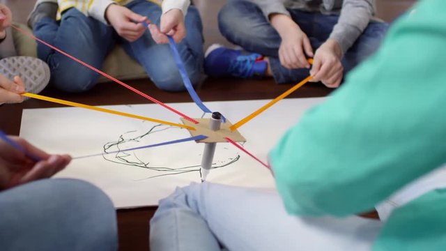 Tilting up close-up shot of group of kids sitting in circle and trying to draw something together on paper with one marker by pulling it in different directions with coloured ribbons