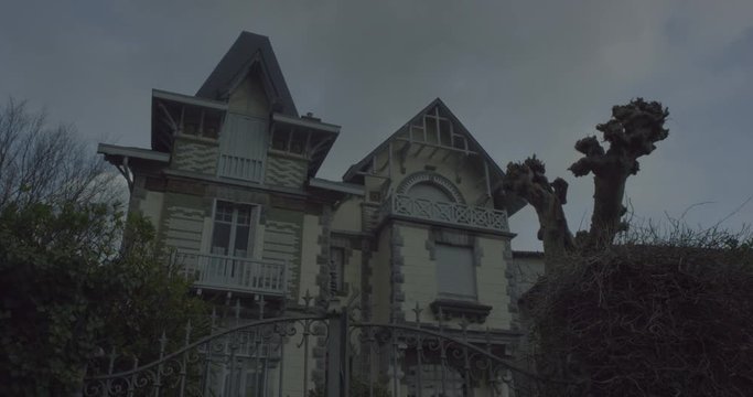 Establishing shot with tilt up of a haunted house in a creepy halloween atmosphere with an imposing old gate and a dead tree and green nature on the sides. Cloudy grey sky. Shot on RED EPIC DRAGON 6K.