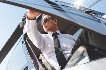 handsome Pilot in sunglasses and formal wear opening door while sitting in helicopter cabin