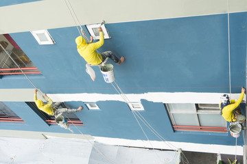Painter hanging on roll, painting color on wall. Facade builder with roller brush, working on high building construction. House exterior contractor commercial, safety worker with lift rope belt.