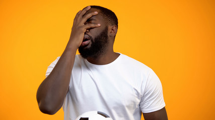 African-American man with soccer ball watching match, showing face palm sign