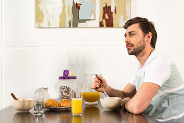 Fototapeta na wymiar bearded man sitting at table during breakfast and dreamy looking away in kitchen