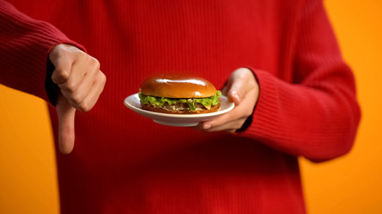 Woman with burger on plate showing thumbs down, fast food obesity, health care