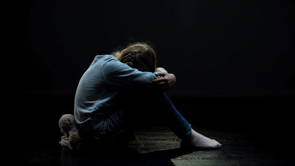 Upset little girl with teddy bear crying in dark abandoned room, loneliness