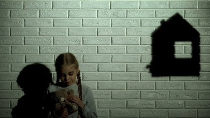 Little girl sadly looking on house shadow and hugging teddy bear, homeless