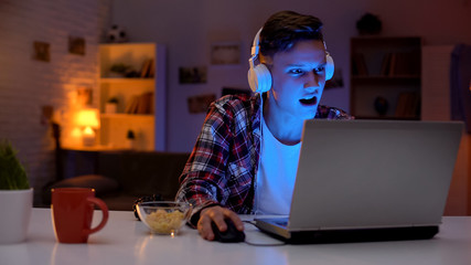 Overemotional teen playing computer game on laptop and eating snacks, addiction