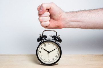 A hand beats a fist on a retro black alarm clock on a wooden table on a white background. Good...