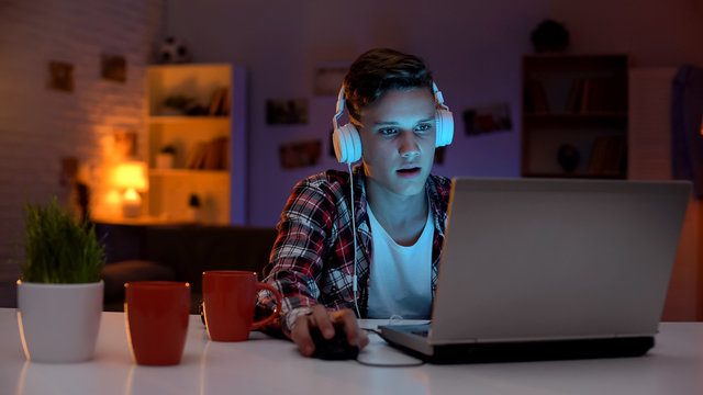 Addicted teenager playing computer games on laptop, lot of cups standing on side