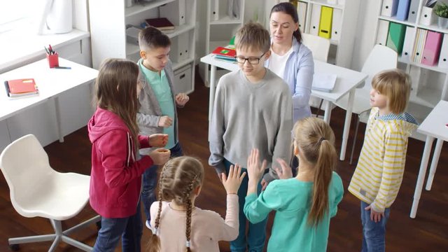 Top shot of casually dressed 13-year-old boy with glasses standing in middle of circle with other kids and woman, who are catching him as he falls towards them with closed eyes