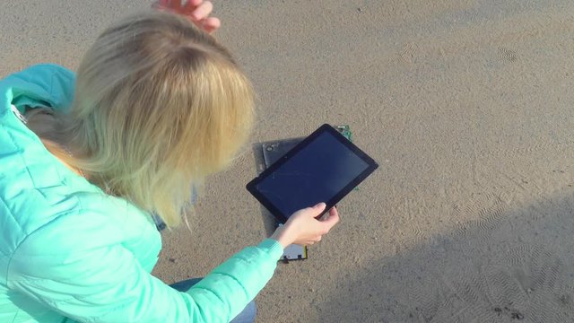 Young beautiful woman, squatting over a tablet shattered into smithereens. The phone is on the pavement, parts are lying around. She holds her head and worries.
