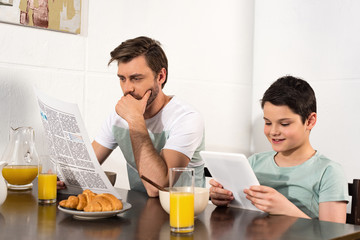 Fototapeta na wymiar pensive father reading newspaper and son using digital tablet during breakfast