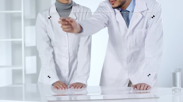 Lockdown of female and male doctors standing together at hi-tech desktop and swiping on glass multi-touch display. Video suitable for adding AR graphics