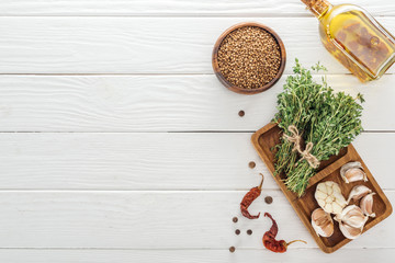 top view of black pepper, dried chili peppers, coriander in bowl near oil in bottle, board with garlic cloves and thyme on white wooden table