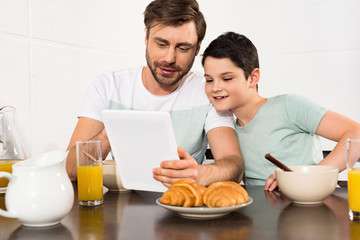 Obraz na płótnie Canvas smiling dad and son using digital tablet during breakfast