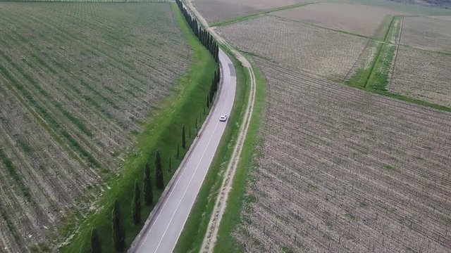 A drone shot of a white Lamborghini Urus, driving on a Tuscan countryside road through a landscape of vineyards and hills.