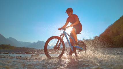 LOW ANGLE: Fit man rides an e-bike in the shallow river and splashes water. - obrazy, fototapety, plakaty