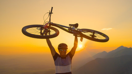 CLOSE UP Man lifts his bicycle above head at sunset after a mountain biking trip