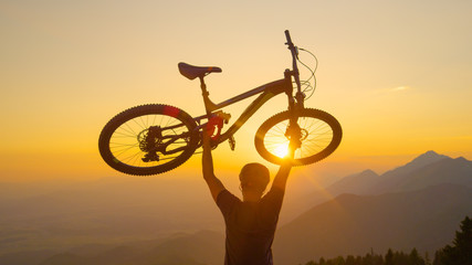Fototapeta na wymiar LENS FLARE: Excited man lifts his bike above his head after a fun bike ride.