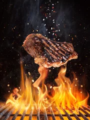 Tuinposter Tasty beef steaks flying above cast iron grate with fire flames. Freeze motion barbecue concept. © Lukas Gojda