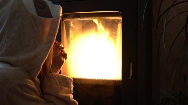 Lonely girl with woolen hood sitting in front of the burning fireplace in the dark. Woman warming up her hands slowly after turning back home from outdoors. Girl rubbing frozen hands at home