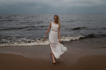 Walking Beautiful young blonde woman beach nymph in white dress near sea with waves during a dull gloomy weather with stormy wind and rain