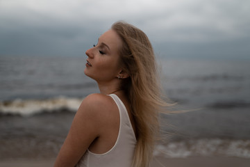 Fototapeta na wymiar Close up portrait of Beautiful young blonde woman beach nymph in white dress near sea with waves during a dull gloomy weather with stormy wind and rain