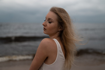 Close up portrait of Beautiful young blonde woman beach nymph in white dress near sea with waves during a dull gloomy weather with stormy wind and rain