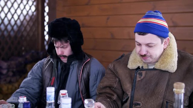 In village two funny men friends dressed in ski hat and brown coat with dark winter hat ear flaps are sitting outside on cold day around table full of food and drinks, talking and eating.