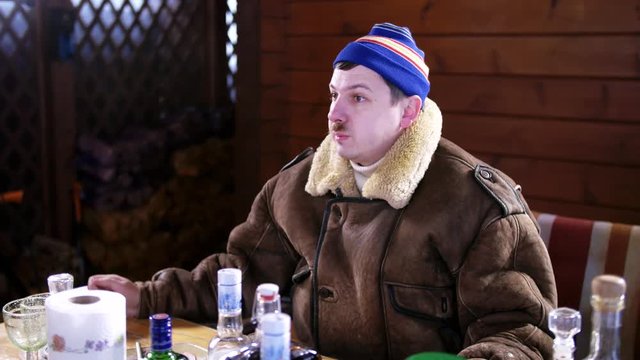Mature funny drunk guy with mustache put on blue cap and thick brown coat suddenly starts twitching and dancing behind table full of food and alcoholic drinks and outside on cold winter day.