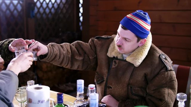 Mature funny drunk male with mustache put on blue cap and thick brown coat gets prepared to drink vodka with friends behind table full of food and alcoholic drinks outside on cold winter day.