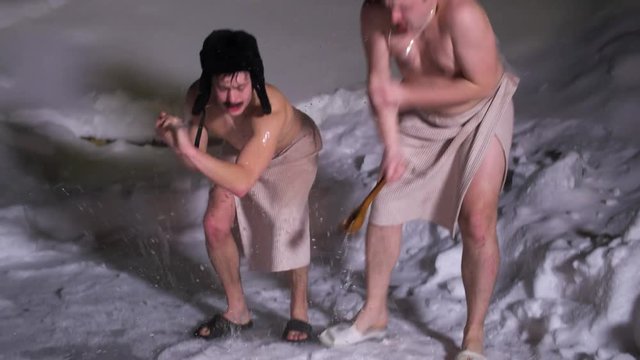 Two funny semi naked men friends wearing blue ski hat and winter hat ear flaps and towel around their waist gathered around huge ice hole on cold day and hit water in it with stick and spoon.